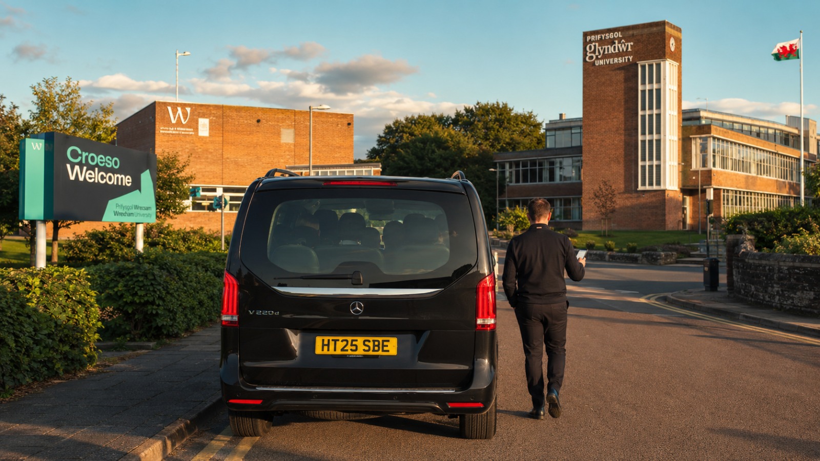 8-seat black minibus departing Wrexham University campus tail-view with student group inside for Manchester Airport